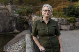 Porträtt kvinna, gråblont hår, glasögon, grön dräkt, ute, träd och grönska i bakgrunden.