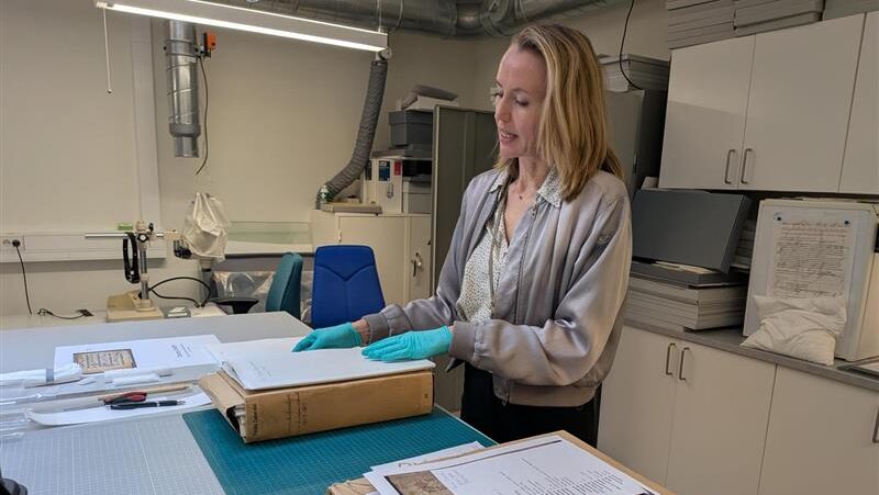 En kvinna med ljust långt hår och turkosa handskar håller i en bunt med pappersark, ovanför en brun arkivbox