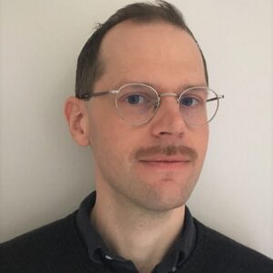 Porträtt, man, kort mörkt hår, mustasch och glasögon, svart skjorta, inne, ljus bakgrund.