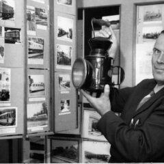 Svartvitt foto där en man håller upp en lådkamera med stor blixt. På väggen en fotoutställning.