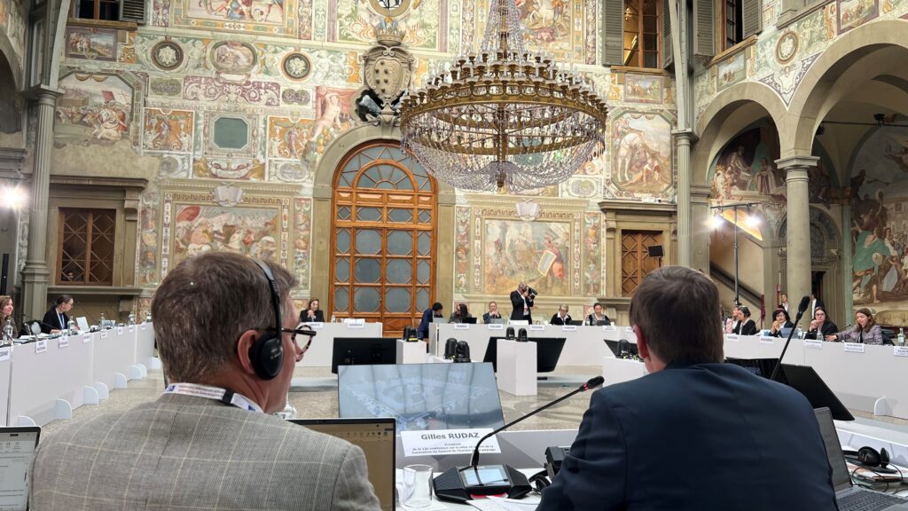 Inne, mötesdeltagare sitter vid bord placerade i cirkelform i en rikt utsmyckad lokal med högt i tak och kristallkronor.