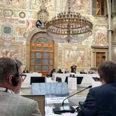 Inne, mötesdeltagare sitter vid bord placerade i cirkelform i en rikt utsmyckad lokal med högt i tak och kristallkronor.