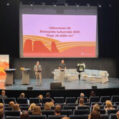 På en stor scen står en man och en kvinna. Scenen är möblerad med ståbord, soffor, blommor och en rollup. I bakgrunden hänger en bildskärm med en välkommentext och en illustration på. I förgrunden sitter publik och tittar uppmärksamt mot talarna.