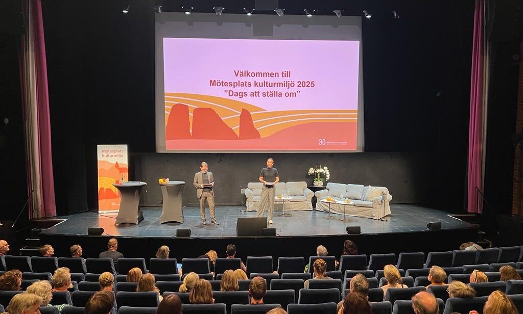 På en stor scen står en man och en kvinna. Scenen är möblerad med ståbord, soffor, blommor och en rollup. I bakgrunden hänger en bildskärm med en välkommentext och en illustration på. I förgrunden sitter publik och tittar uppmärksamt mot talarna.