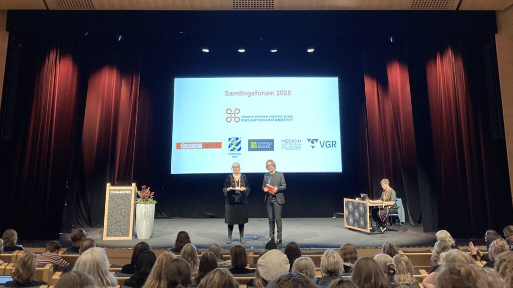 Två personer står på scen framför en duk med texten ”Samlingsforum 2025” och flera logotyper, medan publiken sitter i salongen.