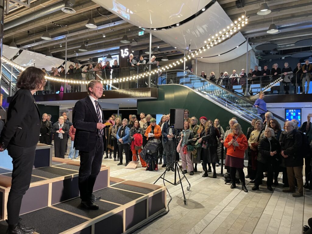 Två personer står på en scen och talar inför en publik