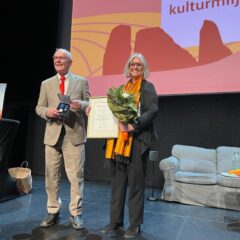 En kvinna med en man på en scen med rödgul skylt bakom. Delar ut pris med blommor och diplom.