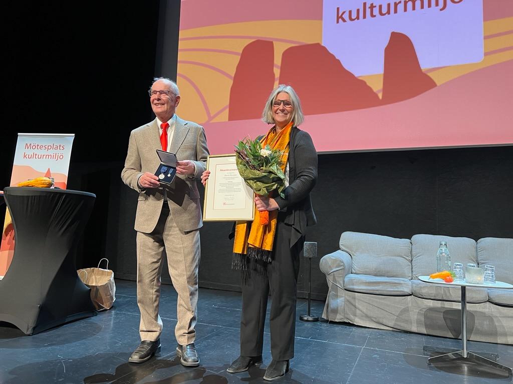 En kvinna med en man på en scen med rödgul skylt bakom. Delar ut pris med blommor och diplom.