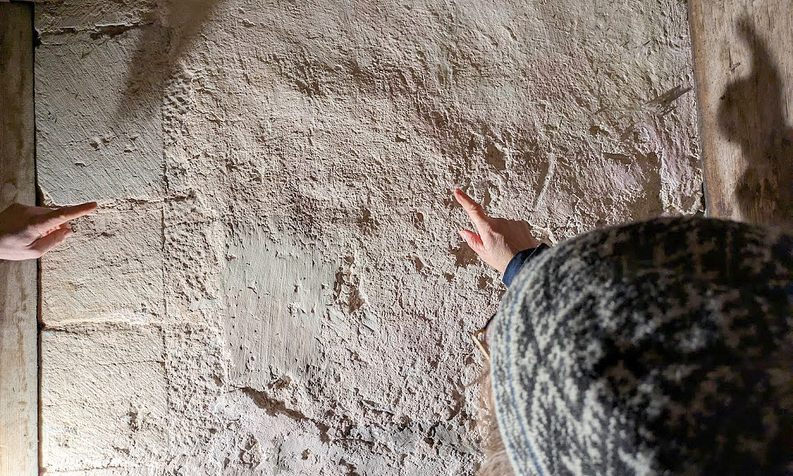 Två personer pekar på märken eller texturer på en gammal stenmur inomhus.