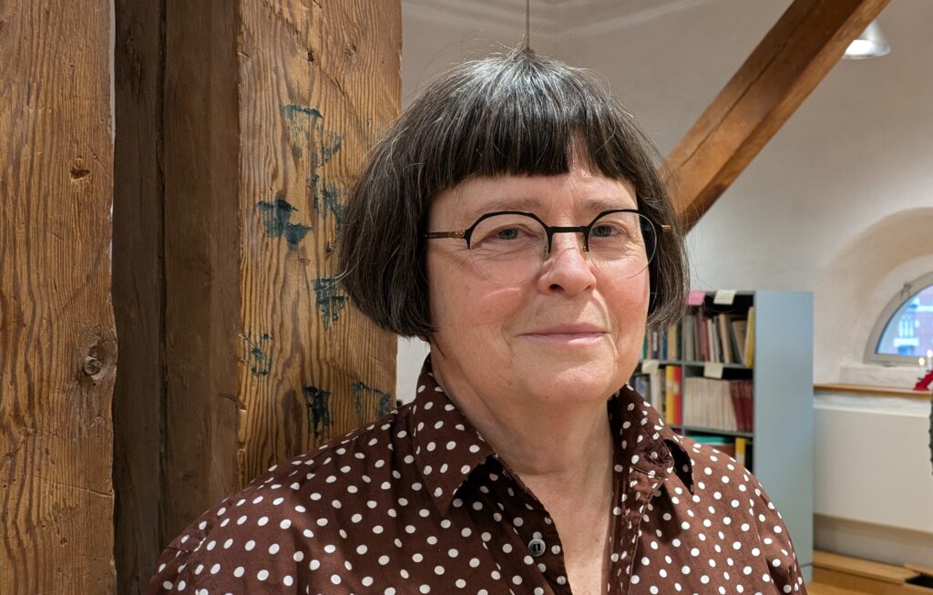 Mörkhårig kvinna med glasögon och prickig blus står framför en trävägg.