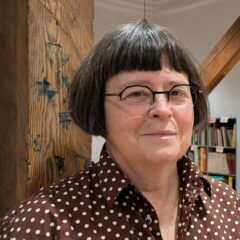 Mörkhårig kvinna med glasögon och prickig blus står framför en trävägg.