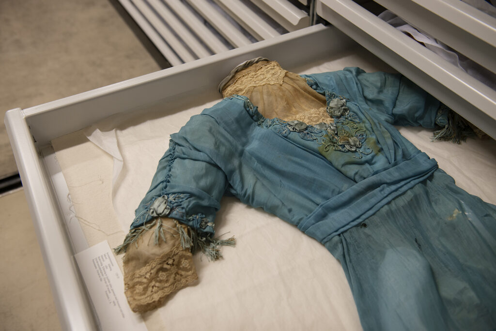 Blå antik klänning med spetsdetaljer, placerad i en låda i museets textilmagasin.