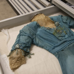 Blå antik klänning med spetsdetaljer, placerad i en låda i museets textilmagasin.