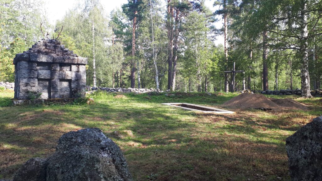 Skogsglänta med gräs och träd. En gammal stenformation med renhorn på toppen och en nygrävd grav med träkors.