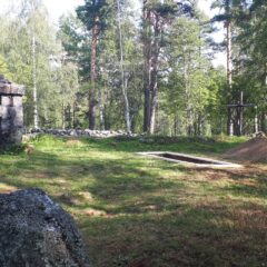 Skogsglänta med gräs och träd. En gammal stenformation med renhorn på toppen och en nygrävd grav med träkors.