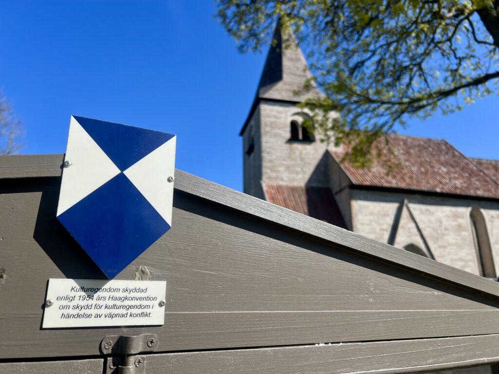 Pilformad skylt, i blått och vitt på stolpe, som pekar mot infoskylt om vad skyddet innebär. I bakgrunden en kyrka.