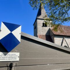 Pilformad skylt, i blått och vitt på stolpe, som pekar mot infoskylt om vad skyddet innebär. I bakgrunden en kyrka.