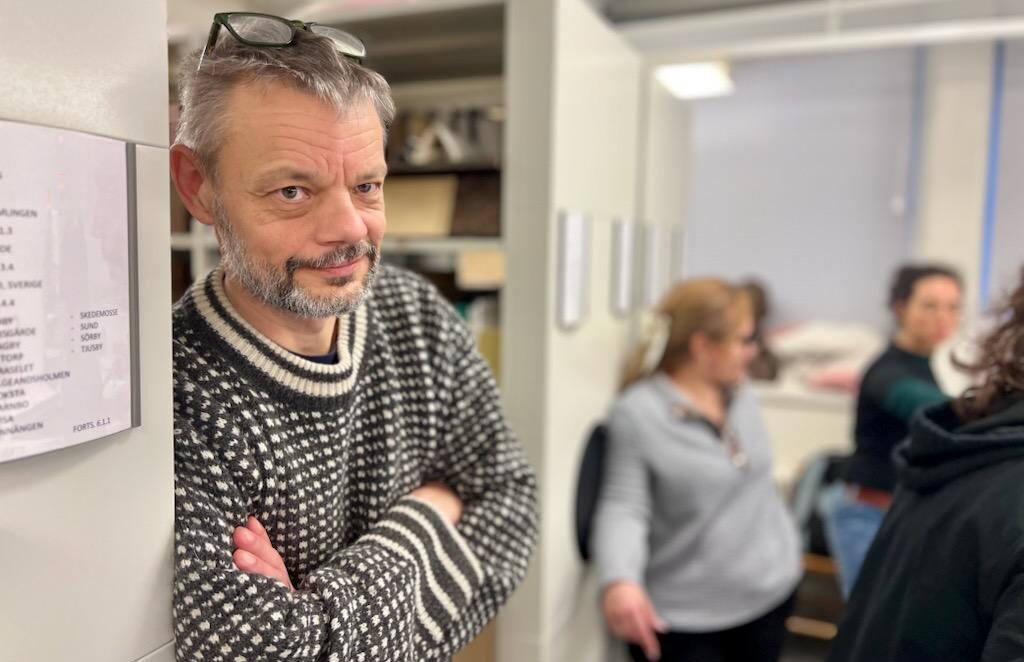 En man med glasögon i pannan, grått hår och skägg, lutar sig mot en arkivhylla i en källarlokal.