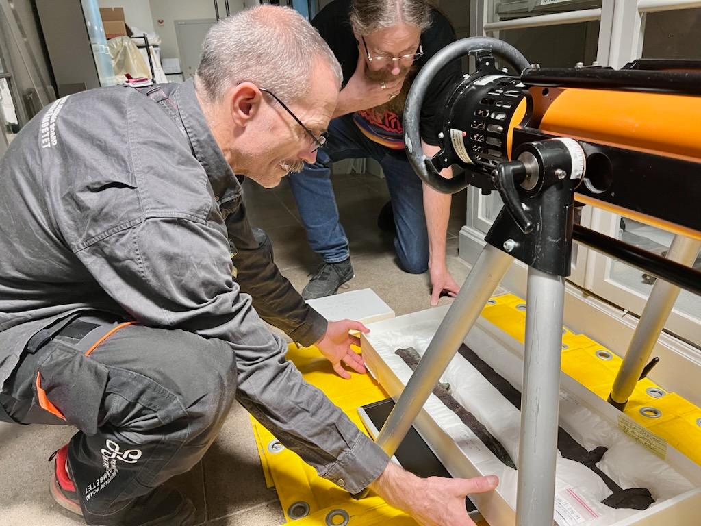 En man sitter på huk på golvet framför ett instrument där han placerar ett rostigt svärd.