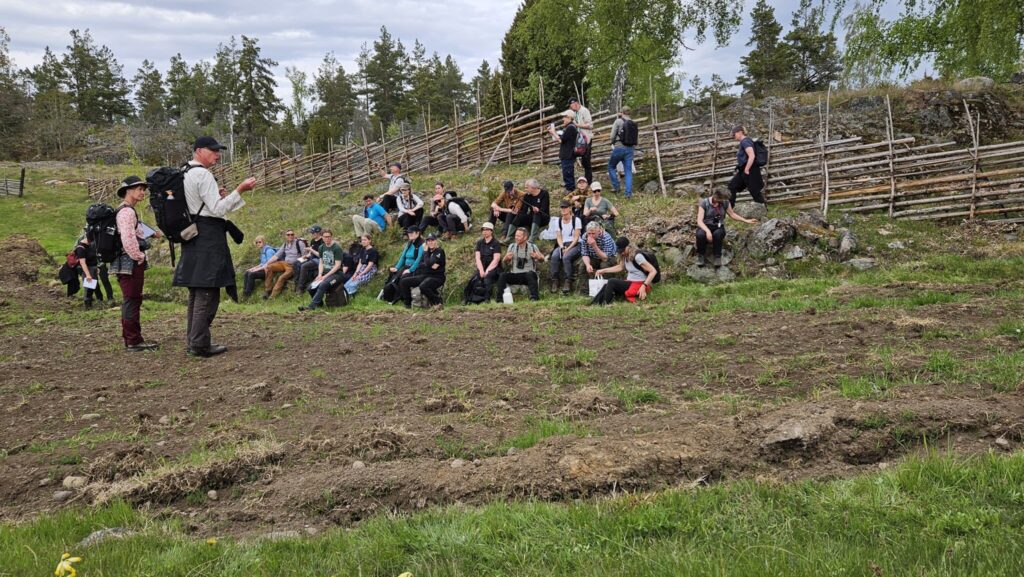 En kursledare står framför ett tiotal deltagare som sitter eller ligger i en lätt lutande backe ovanför en åker.