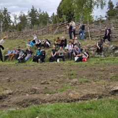 En kursledare står framför ett tiotal deltagare som sitter eller ligger i en lätt lutande backe ovanför en åker.