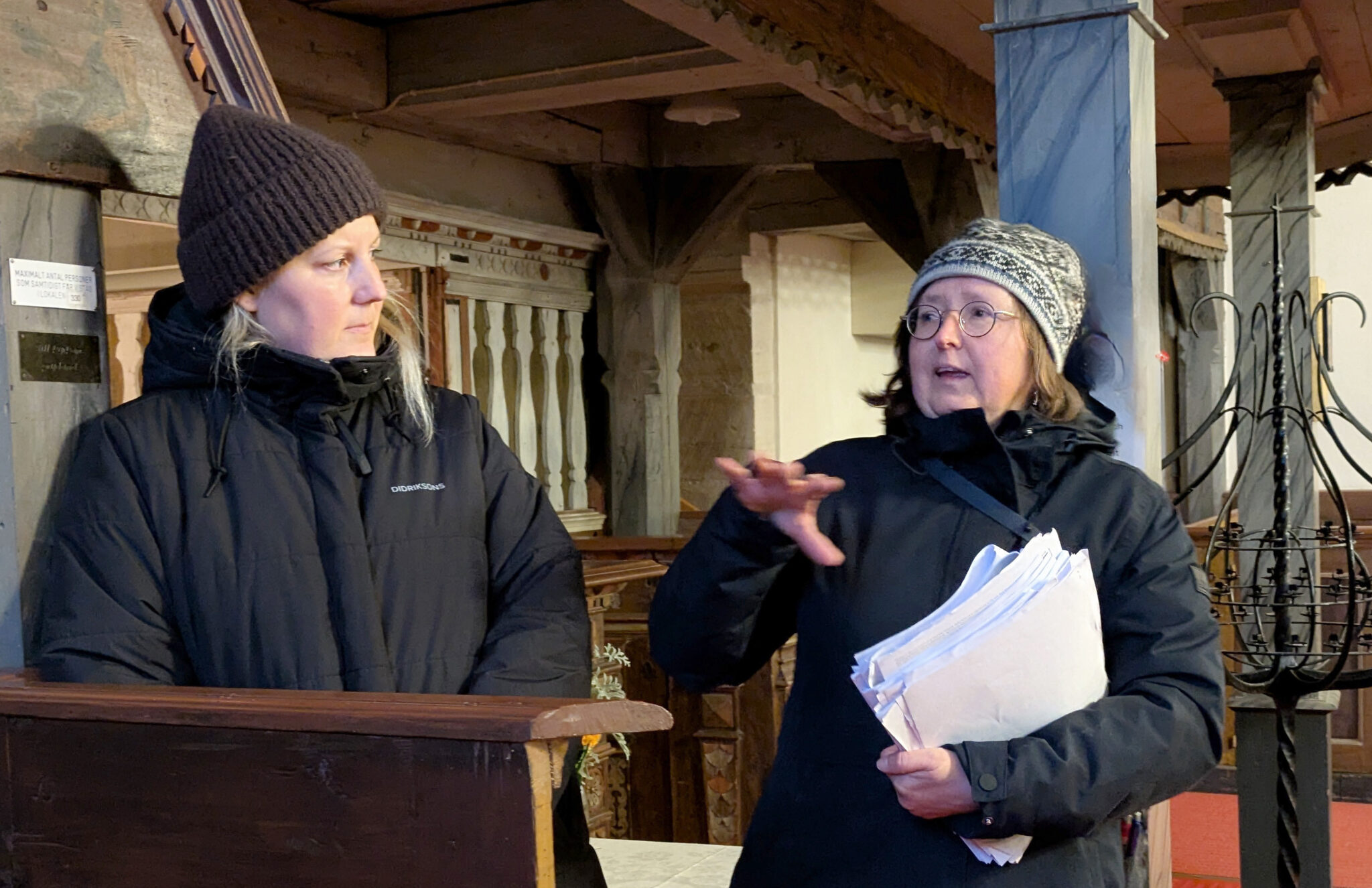 Två personer står inomhus och diskuterar, en håller papper och gestikulerar; båda i vinterkläder.