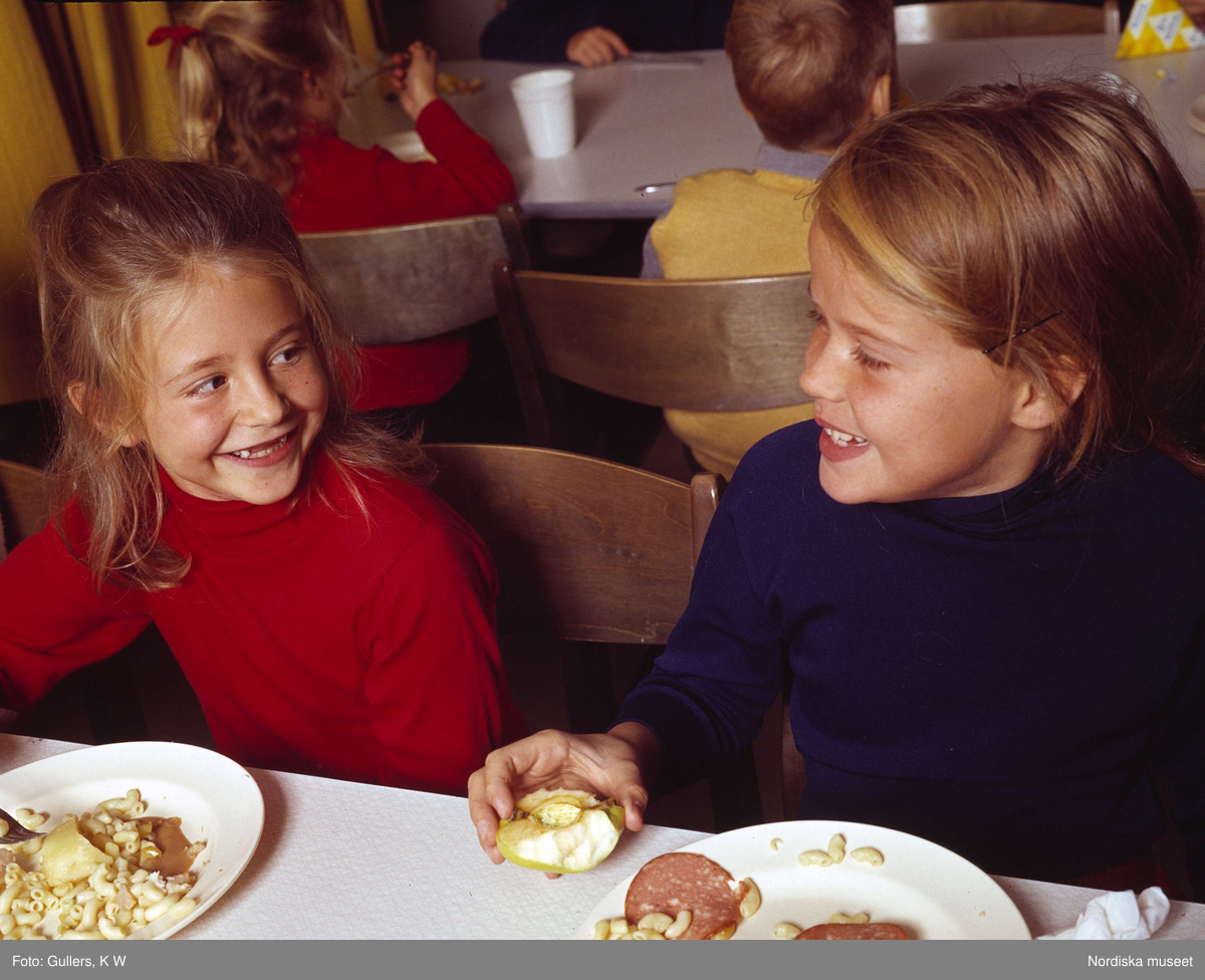 Två blonda flickor i 10-årsåldern med röd och blå tröja sitter vi ett matbord och tittar på varandra