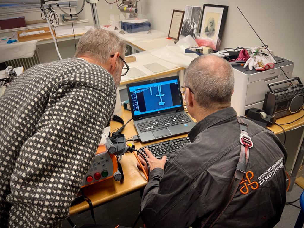 Två män med ryggen mot kameran granskar en bild i en datorskärm.