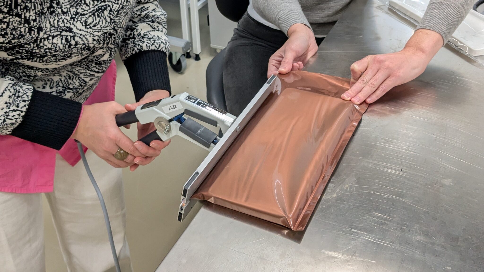 Två händer håller i ett tångliknande verktyg och två andra händer håller den kopparfärgade plastpåsen.