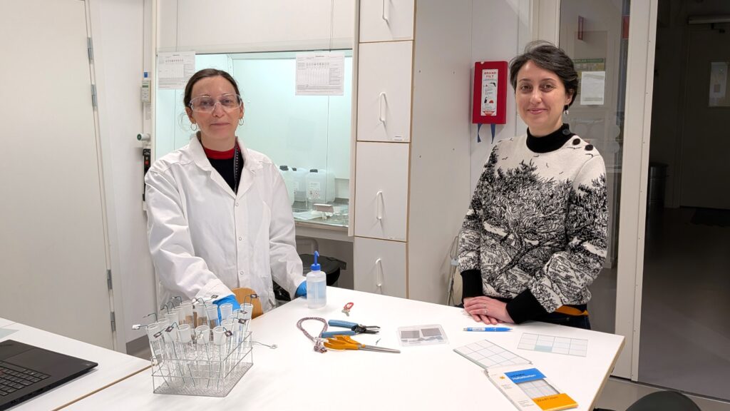 Två mörkhåriga kvinnor står framför ett bord i ett laboratorium. Den en är klädd i labbrock och labbglasögon, och den andra i stickad tröja.