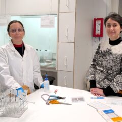 Två mörkhåriga kvinnor står framför ett bord i ett laboratorium. Den en är klädd i labbrock och labbglasögon, och den andra i stickad tröja.