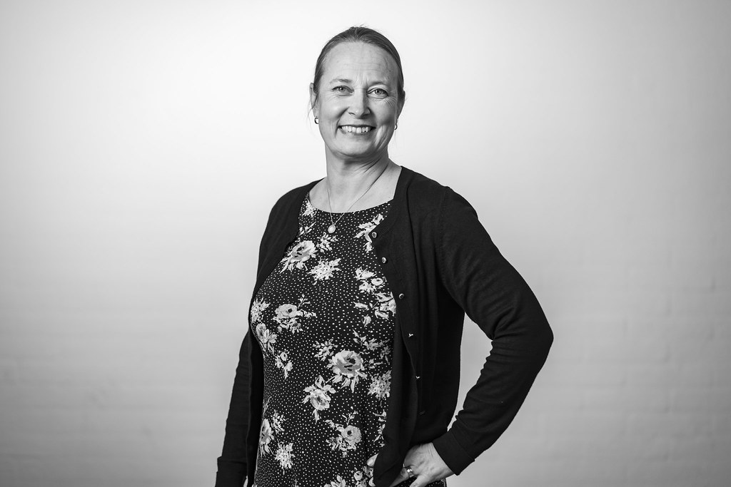 Porträtt av person i blommig topp och mörk kofta, står mot ljus bakgrund och ler mot kameran