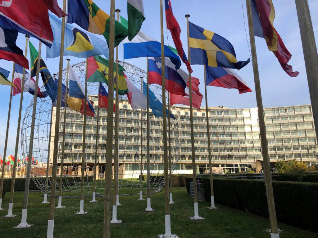 Framför en vit halvmåneformad byggnad med många våningar står ståtliga flaggstänger med all världens flaggor.