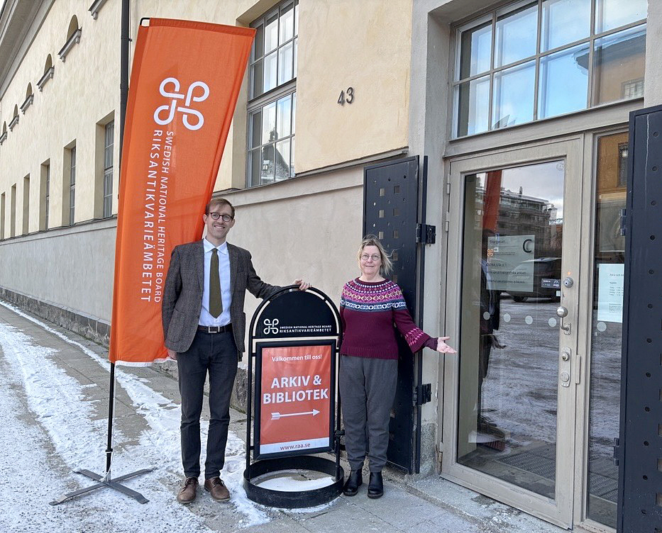 Två personer står utanför en byggnad med glasdörrar och adressnumret 43. Bredvid dem finns en hög orange flagga och en fristående skylt som visar vägen till “Arkiv & Bibliotek” vid Riksantikvarieämbetet. Markerna är täckta av snö och båda personerna poserar välkomnande bredvid skylten.