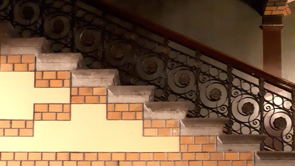 En trappa med utsirat räcke leder från höger upp mot vänster. Dekor av tegel och gul puts på sidan.
