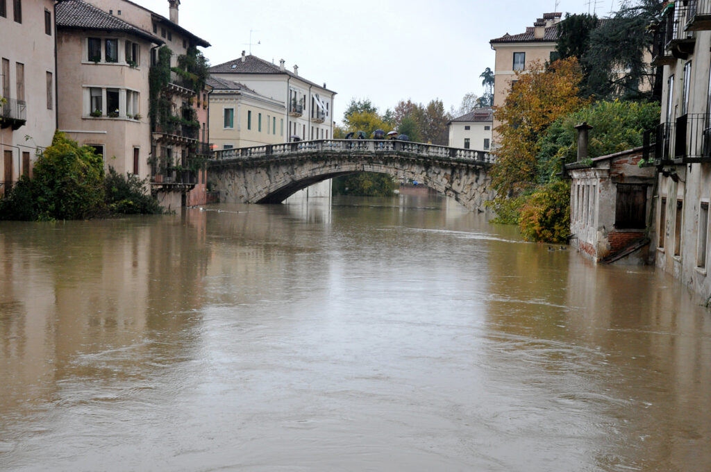 En stenbro sträcker sig över en flod som svämmat över, med vatten som nästan når upp till byggnaderna längs kajen en mulen höstdag.