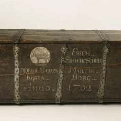 Kraftig järnbeslagen kista av ek med plant lock och mot bottnen sluttande sidor. Profilerad nederkant runt kistans botten. Kraftiga smidda bärhandtag. Mycket tungt lock. På framsidan med originalmålad text i versaler i vit oljefärg.