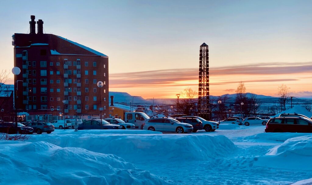 Snötäckt parkering med bilar framför ett högt tegelhus och ett torn, solnedgång över fjäll i bakgrunden.