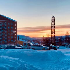 Snötäckt parkering med bilar framför ett högt tegelhus och ett torn, solnedgång över fjäll i bakgrunden.