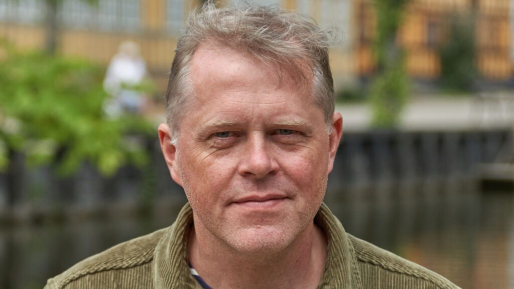 Porträttbild på ljushårig man i grön skorta med randig t-shirt under.