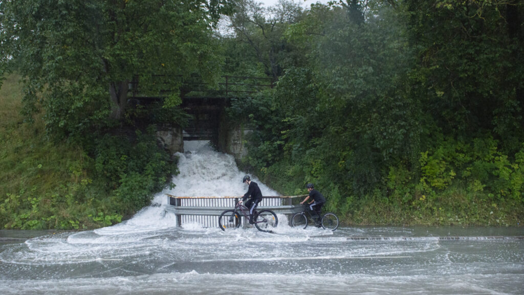 Två personer cyklar över en översvämmad å med en palissadbro över.