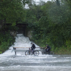 Två personer cyklar över en översvämmad å med en palissadbro över.