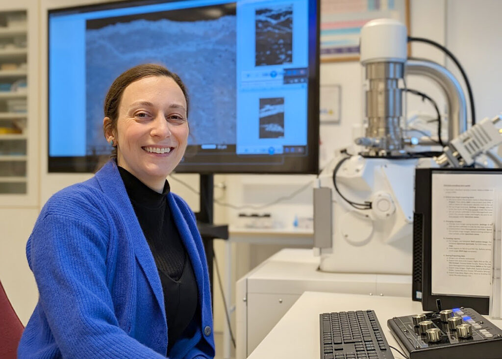 En person i blå kofta sitter vid ett skrivbord i ett labb bredvid ett elektronmikroskop, med en stor skärm bakom sig som visar en närbild av ett materialprov.
