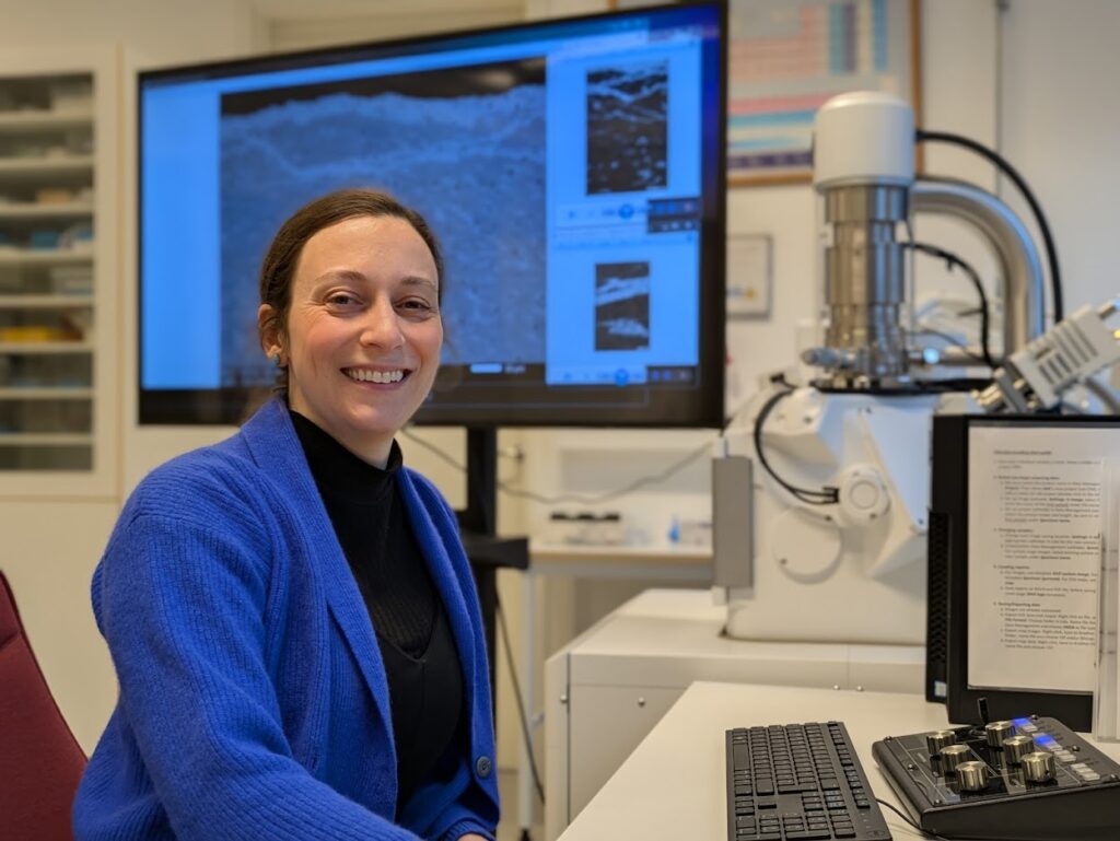 En person i blå kofta sitter vid ett skrivbord i ett labb bredvid ett elektronmikroskop, med en stor skärm bakom sig som visar en närbild av ett materialprov.