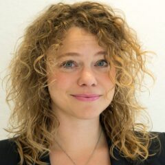 Kvinna med leende mun och rödaktigt lockigt halvlångt hår, iklädd svart kavaj, och ler mot kameran.