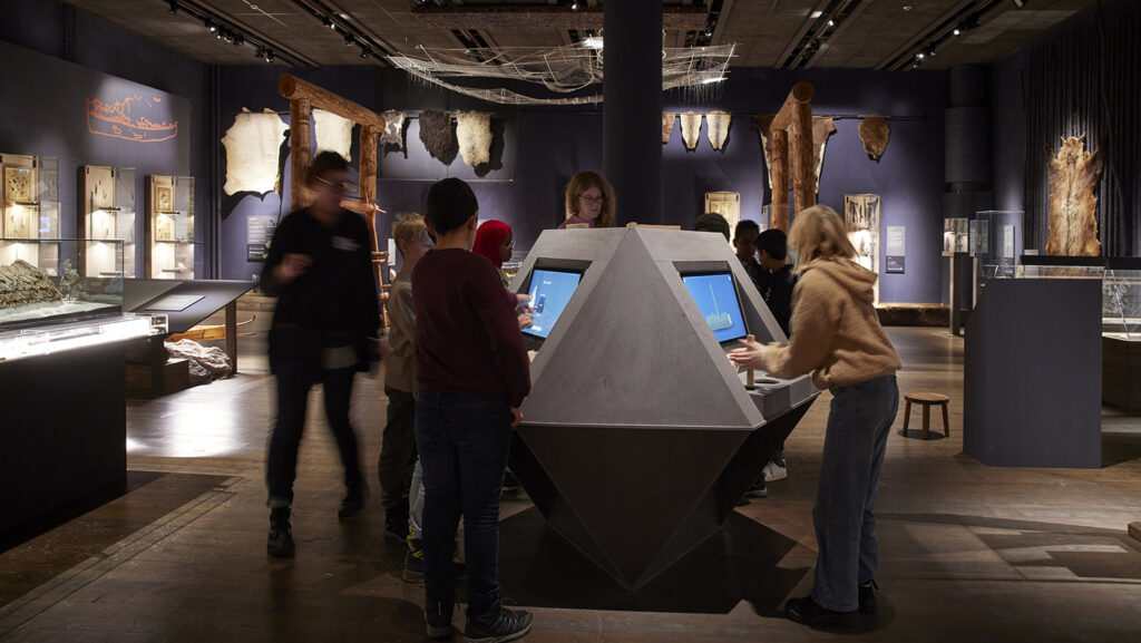 Besökare, varav några barn, står runt en trekantig monter med två TV-skärmar.