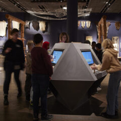 Besökare, varav några barn, står runt en trekantig monter med två TV-skärmar.