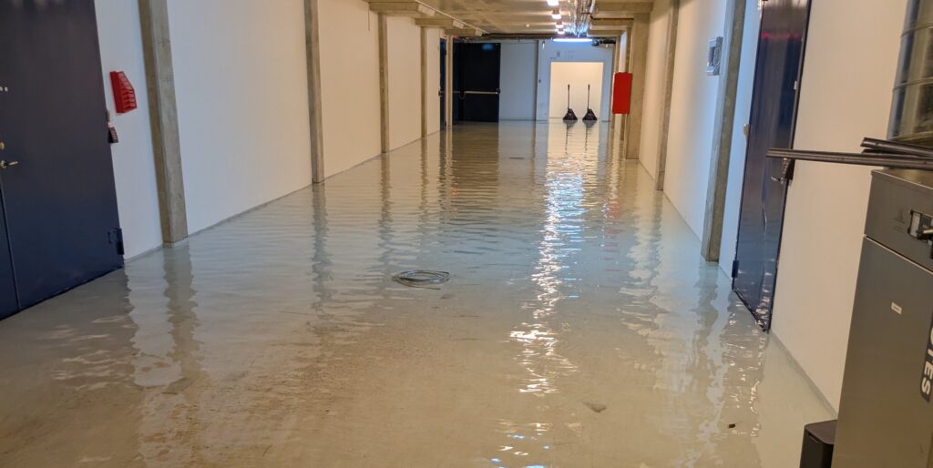 En lång korridor i en byggnad där golvet står helt under vatten. Vattnet täcker hela gången och reflekterar taklamporna. Längs väggarna syns dörrar, brandsläckare och utrustning, och i förgrunden står en robotdammsugare delvis nedsänkt i vattnet.