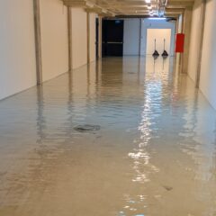 En lång korridor i en byggnad där golvet står helt under vatten. Vattnet täcker hela gången och reflekterar taklamporna. Längs väggarna syns dörrar, brandsläckare och utrustning, och i förgrunden står en robotdammsugare delvis nedsänkt i vattnet.