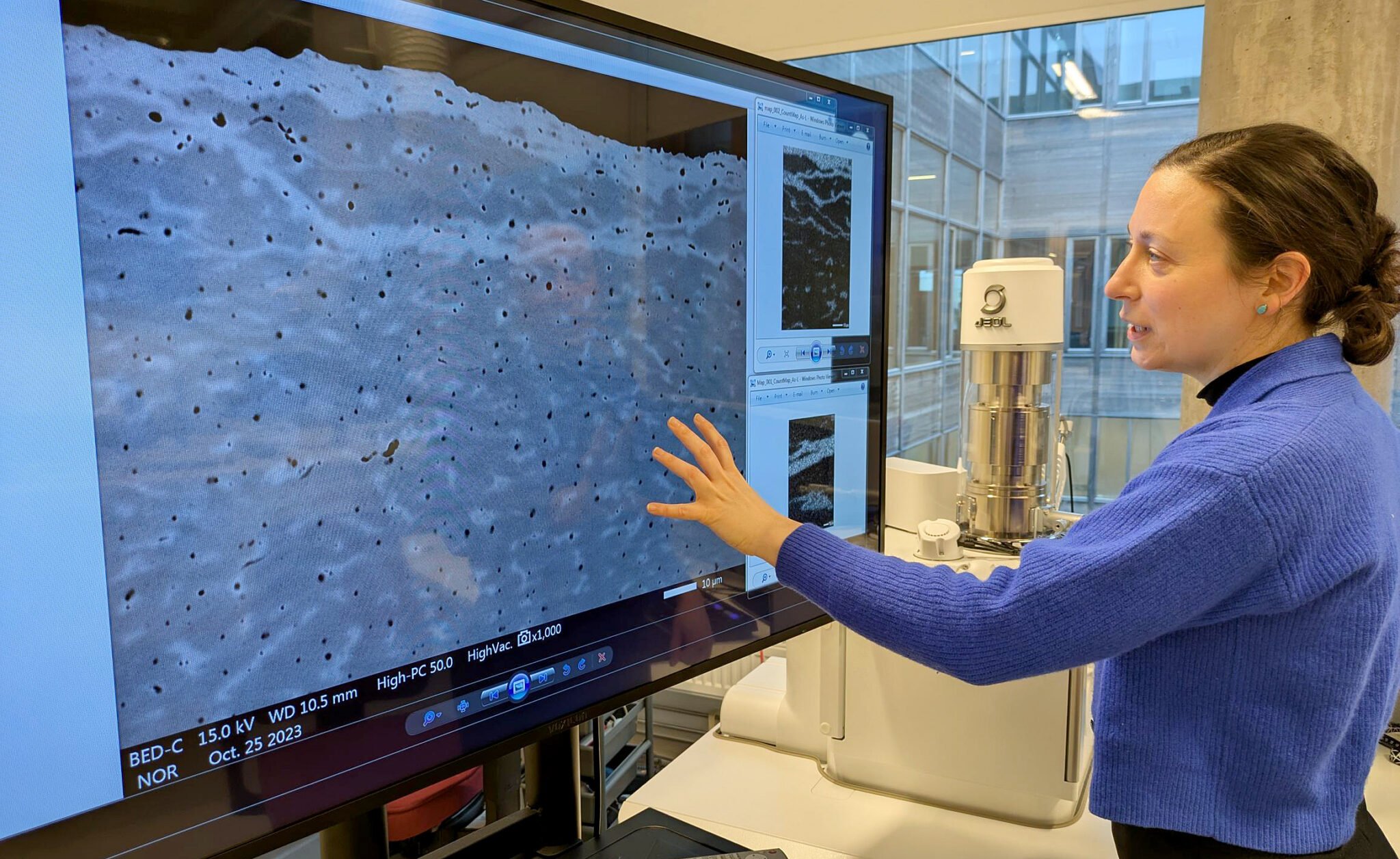 En person i blå kofta står vid en stor skärm och pekar på en förstoring av en materialytas struktur i ett labb med avancerad mikroskoputrustning.